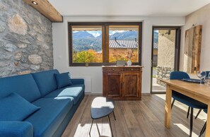 Ein Zimmer mit einer Steinswand, einem blauen Sofa, einem Holzkabinett, einem Esstisch und Bergblick durch ein Fenster.
