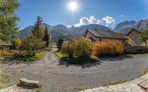 Eine Landstraße führt zu einem Haus mit einem Kiesweg, umgeben von Bäumen und Bergen.
