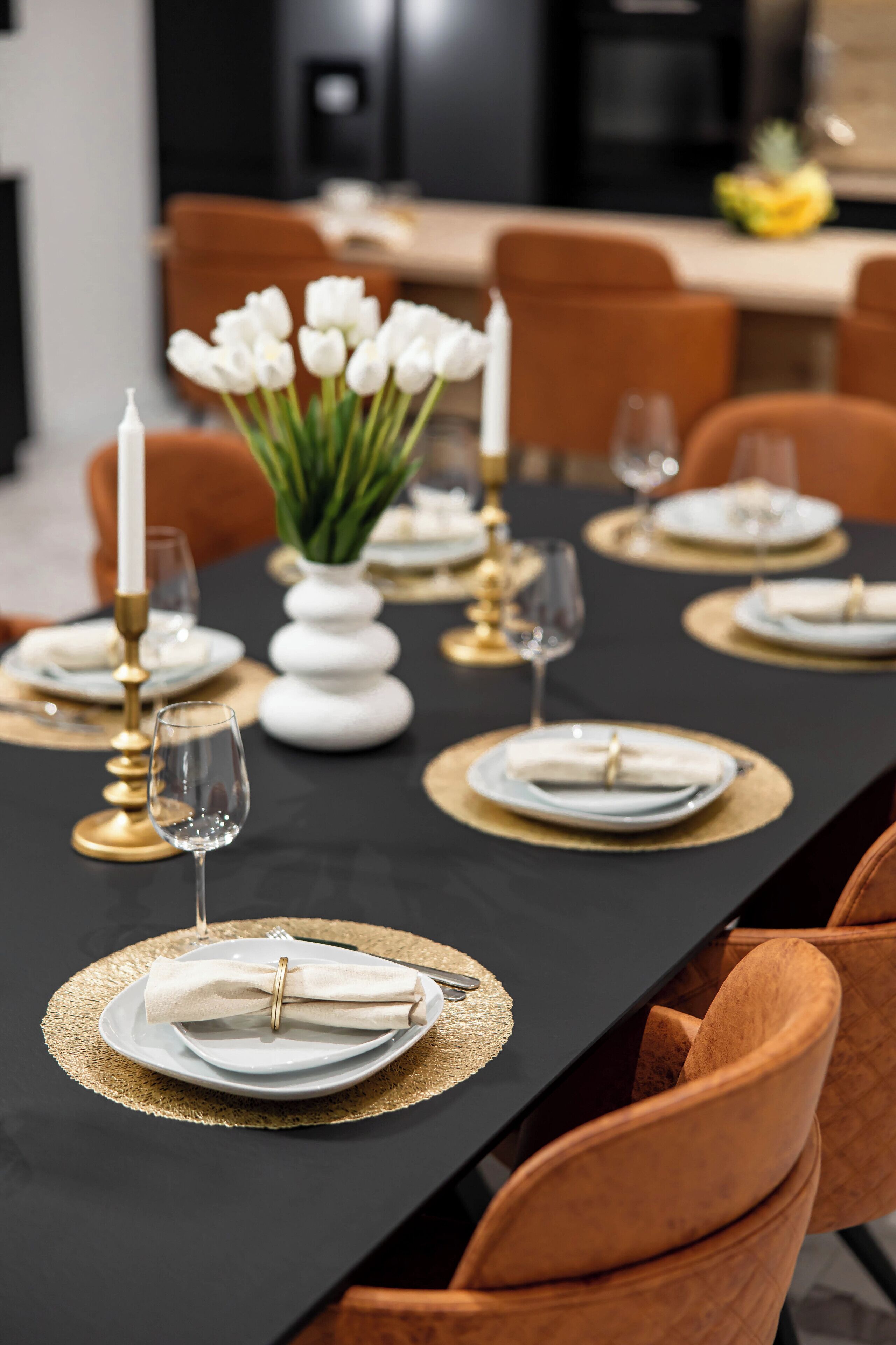 Een eettafel gedekt met gouden placemats, witte borden en gouden kandelaars.