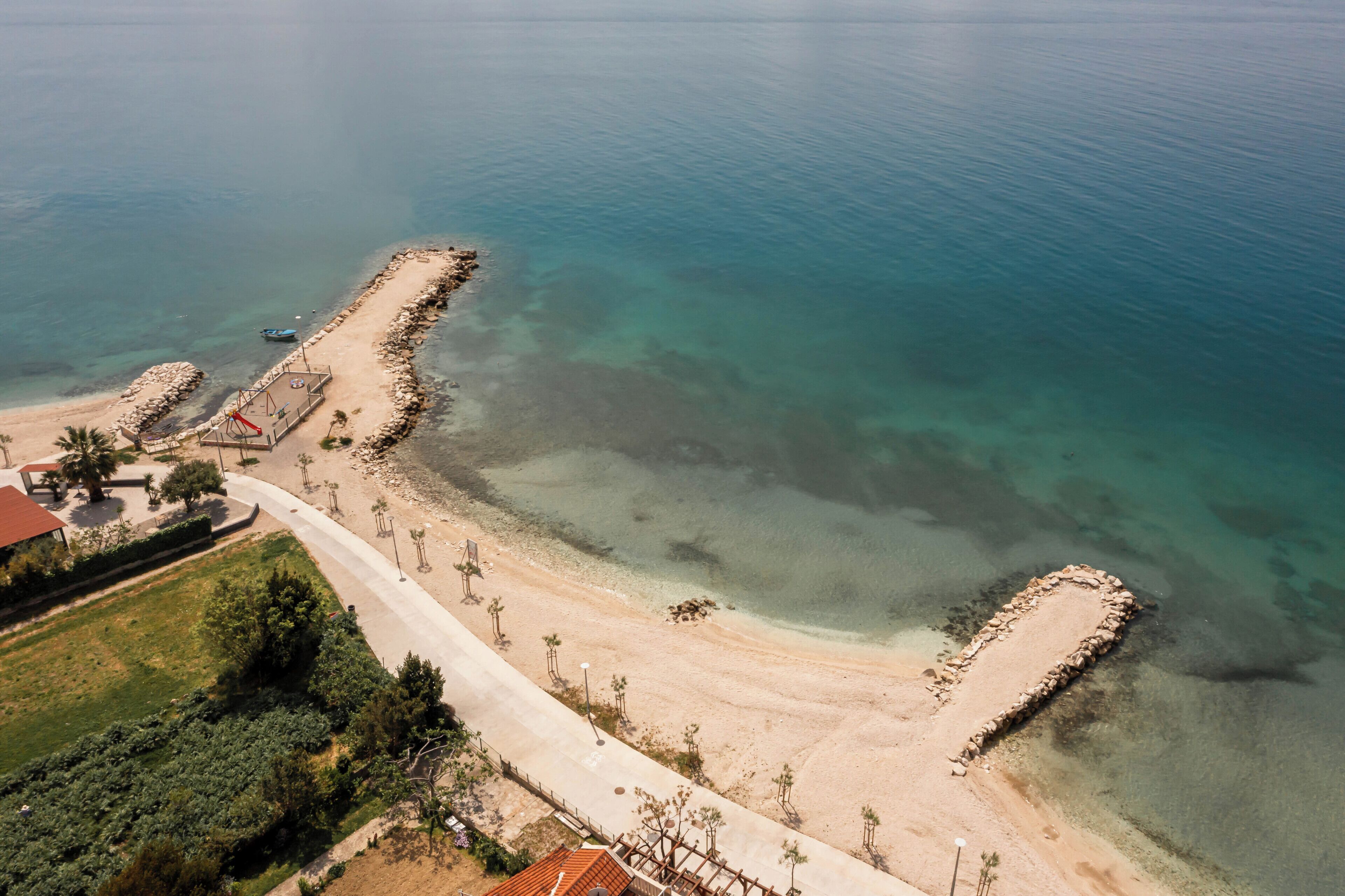An aerial view of a coastal area with a clear beach, a paved walkway, and a small building with a red roof.