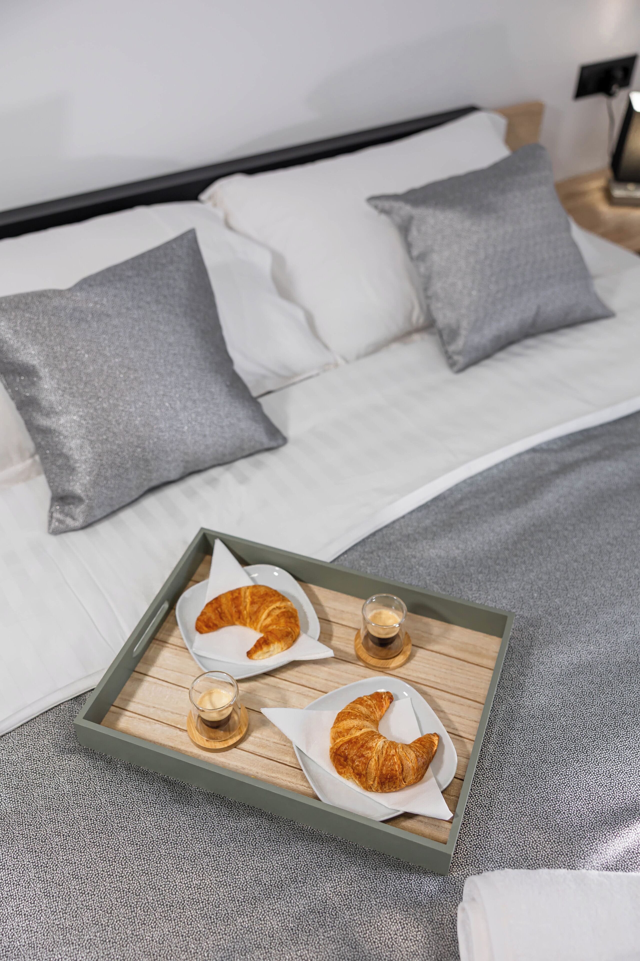 A bed with pillows, a tray with croissants and coffee, and a bedside table with a lamp.