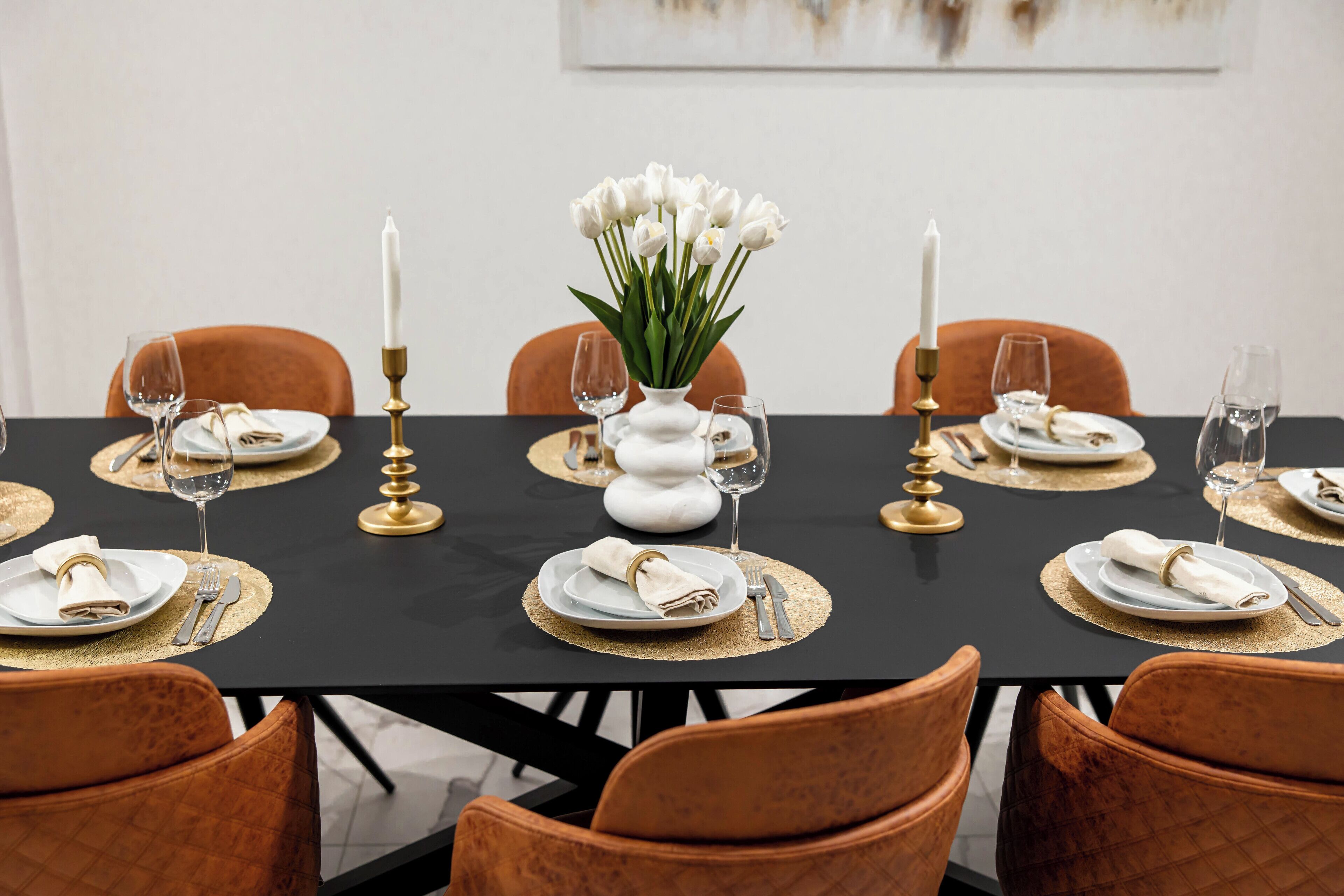 A dining table set with white plates, gold placemats, and a vase of white flowers.