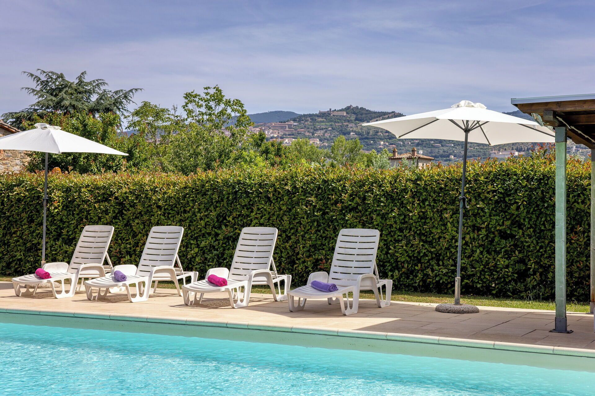 Un espace piscine avec des chaises longues et des parasols, une haie et une vue sur une ville en arrière-plan.