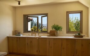 A kitchen with wooden cabinets, a sink, and two windows with views of greenery and a desert landscape. - Los Tilos Estate: Luxury Spanish Estate with Iconic LA Views & Private Pool (Los Angeles)