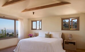A bedroom with a large bed, two bedside tables, a window with a view, and a wooden ceiling beam. - Los Tilos Estate: Luxury Spanish Estate with Iconic LA Views & Private Pool (Los Angeles)