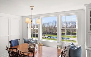 Ein Essbereich mit Holztisch, Stühlen und einer Pendelleuchte. Große Fenster bieten Blick auf einen Garten und ein Schwimmbecken.