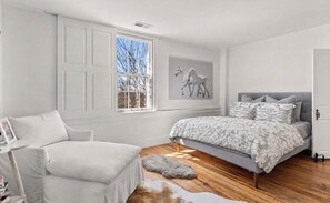Ein Schlafzimmer mit einem Bett, einem Sessel, einem Fenster und einem gerahmten Bild an der Wand.