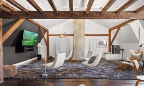 Ein geräumiges Dachzimmer mit freiliegenden Holzbalken, einem großen Flachbildfernseher, weißen Ledersesseln, einem gemusterten Teppich und einem Steinkamin.
