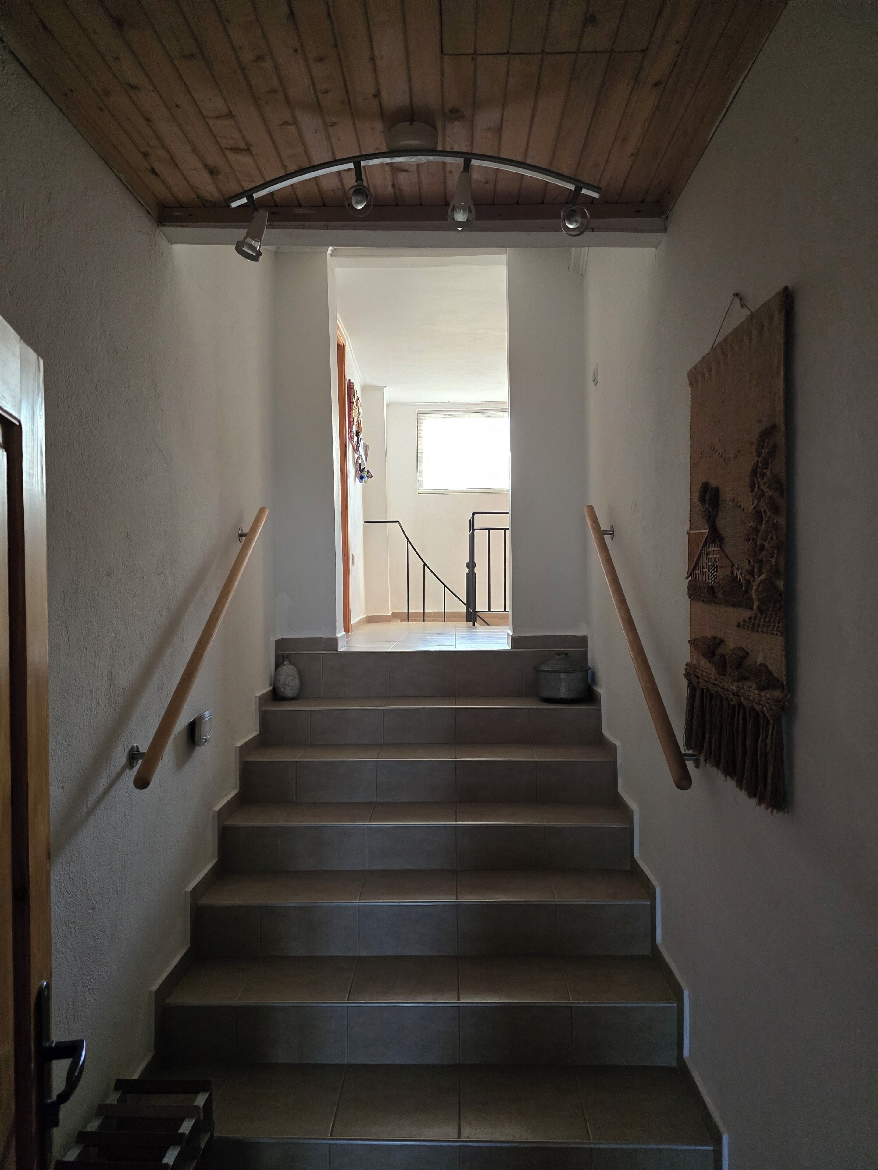 A staircase with wooden handrails, a light fixture above, and a textured wall on the right.
