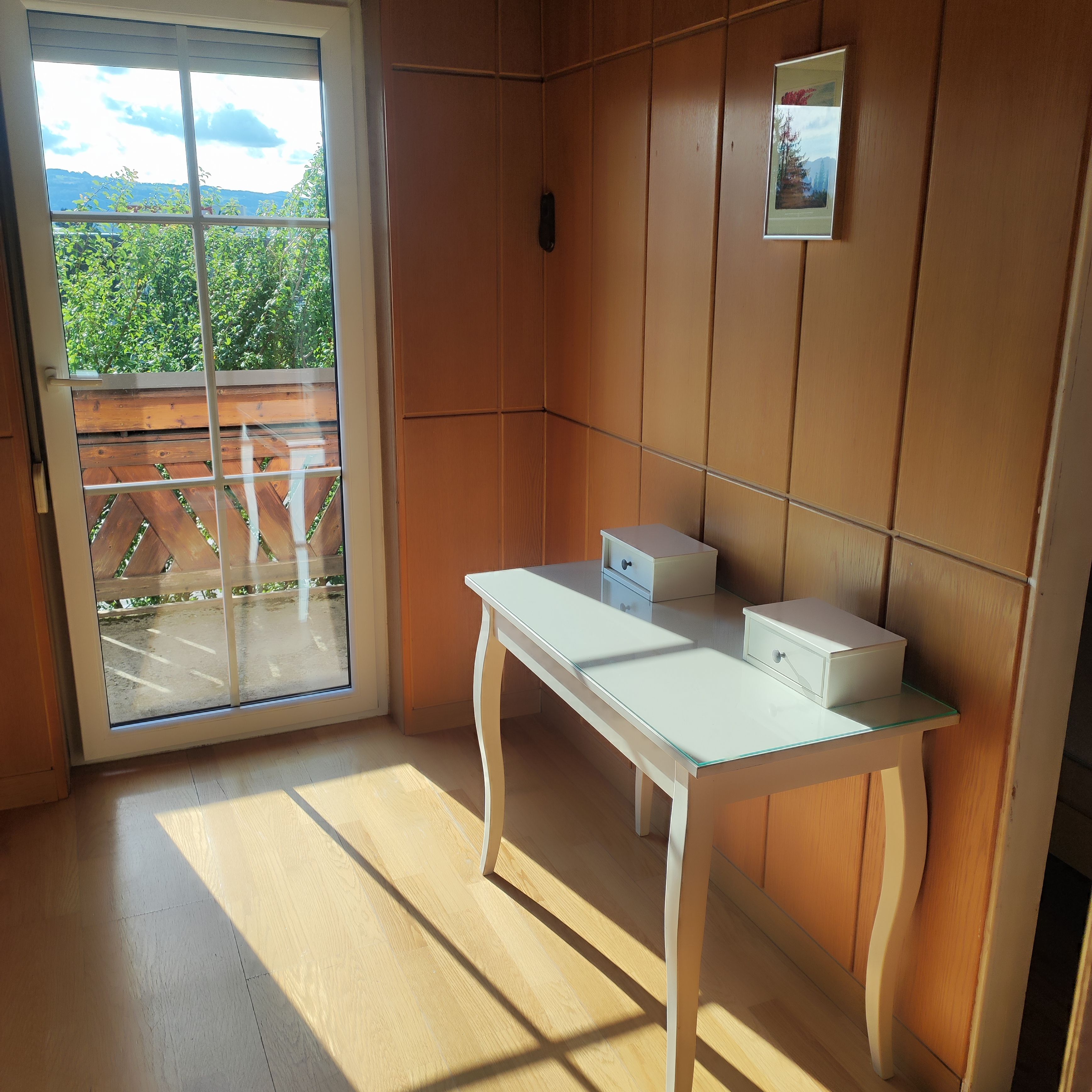 Ein Zimmer mit einer Schiebeglastür, die auf einen Balkon führt, einem weißen Tisch mit Schubladen und einem gerahmten Bild an der Wand.