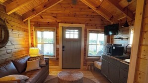 A log cabin interior with a wooden ceiling, a sofa, a round woven ottoman, a kitchen area, and a television. - Tranquil Cabin In Charming Bean Station Tn on Cherokee Lake With Boat Ramp (Bean Station)