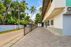 A paved walkway leading to a building with a gated entrance, surrounded by lush greenery and palm trees.