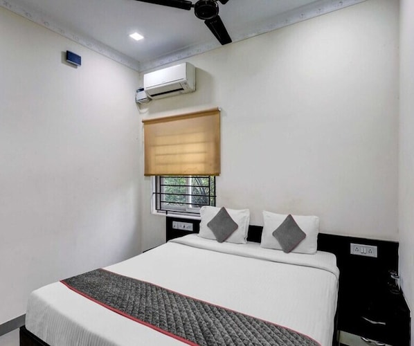 A hotel room with a bed, a ceiling fan, an air conditioning unit, a window with a blind, and a bedside table.