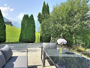 Ein Tisch mit Glasplatte, eine Vase mit Blumen, ein graues Sofa und Blick auf eine gebirgige Landschaft mit ordentlich geschnittenen Hecken.