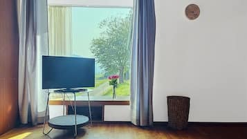A room with a television on a stand, a window with curtains, a clock on the wall, and a trash bin.