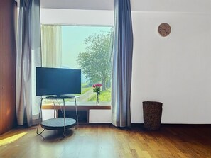 A room with a television on a stand, a window with curtains, a clock on the wall, and a trash bin.