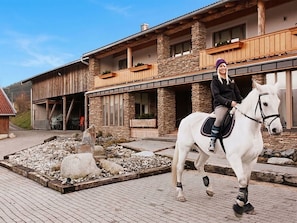 A person riding a white horse in front of a building with stone pillars and a wooden balcony.