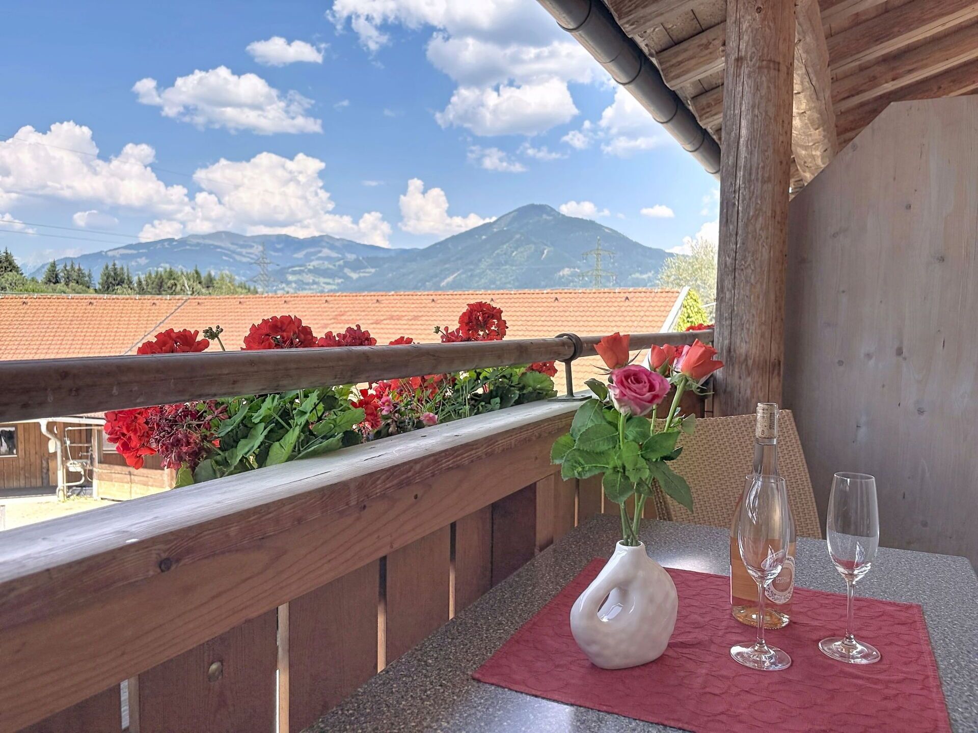 A balcony with a wooden railing, a vase with flowers, and two wine glasses on a table.