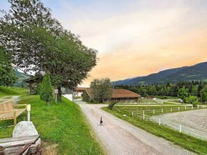 A rural setting with a gravel path leading to a wooden building, surrounded by greenery and mountains in the background.