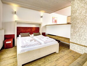Une chambre d’hôtel avec un plancher en bois, un lit recouvert de draps blancs, une tête de lit rouge et un mur en pierre.
