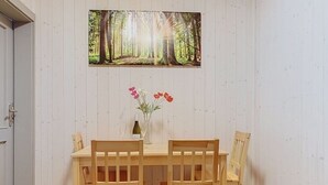 Ruokailutilassa on puinen pöytä ja tuolit, metsäaiheinen maalaus sekä maljakko, jossa on kukkia.