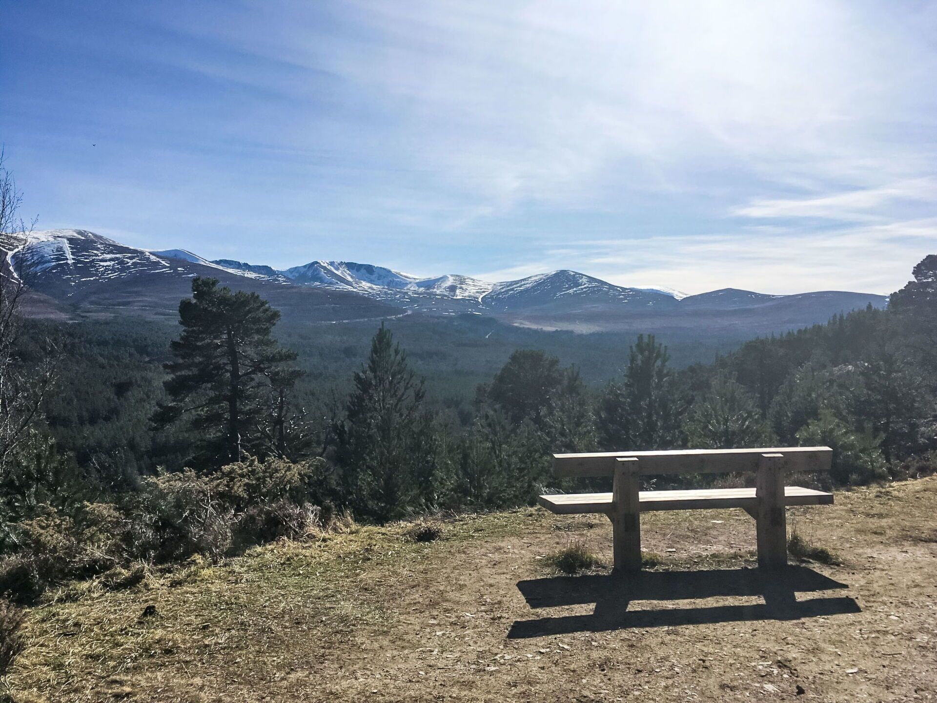 A wooden bench on a clear day overlooking a mountainous landscape with snow-capped peaks.