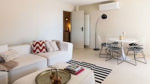 Ein modernes Wohnzimmer mit einem weißen Sofa, einem Esstisch mit Stuhlgestellen aus Draht, einer schwarzen Stehlampe und einem Streifenmuster-Teppich.