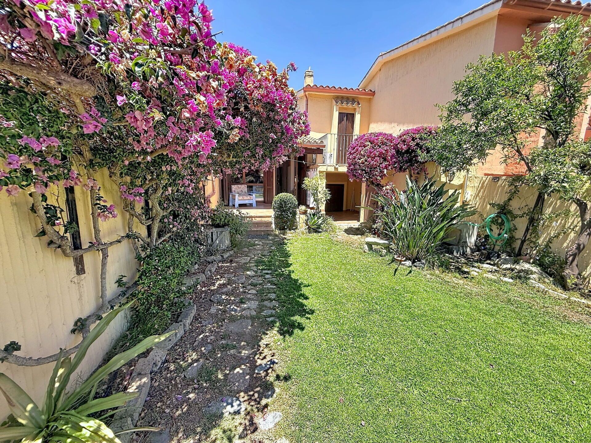 Un patio con un sendero de piedra que conduce a un edificio, rodeado de exuberante follaje y plantas con flores.