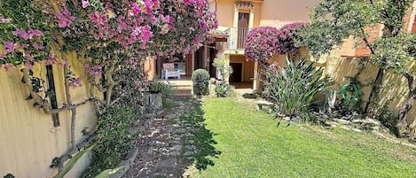 A courtyard with a stone pathway leading to a building, surrounded by lush greenery and flowering plants.