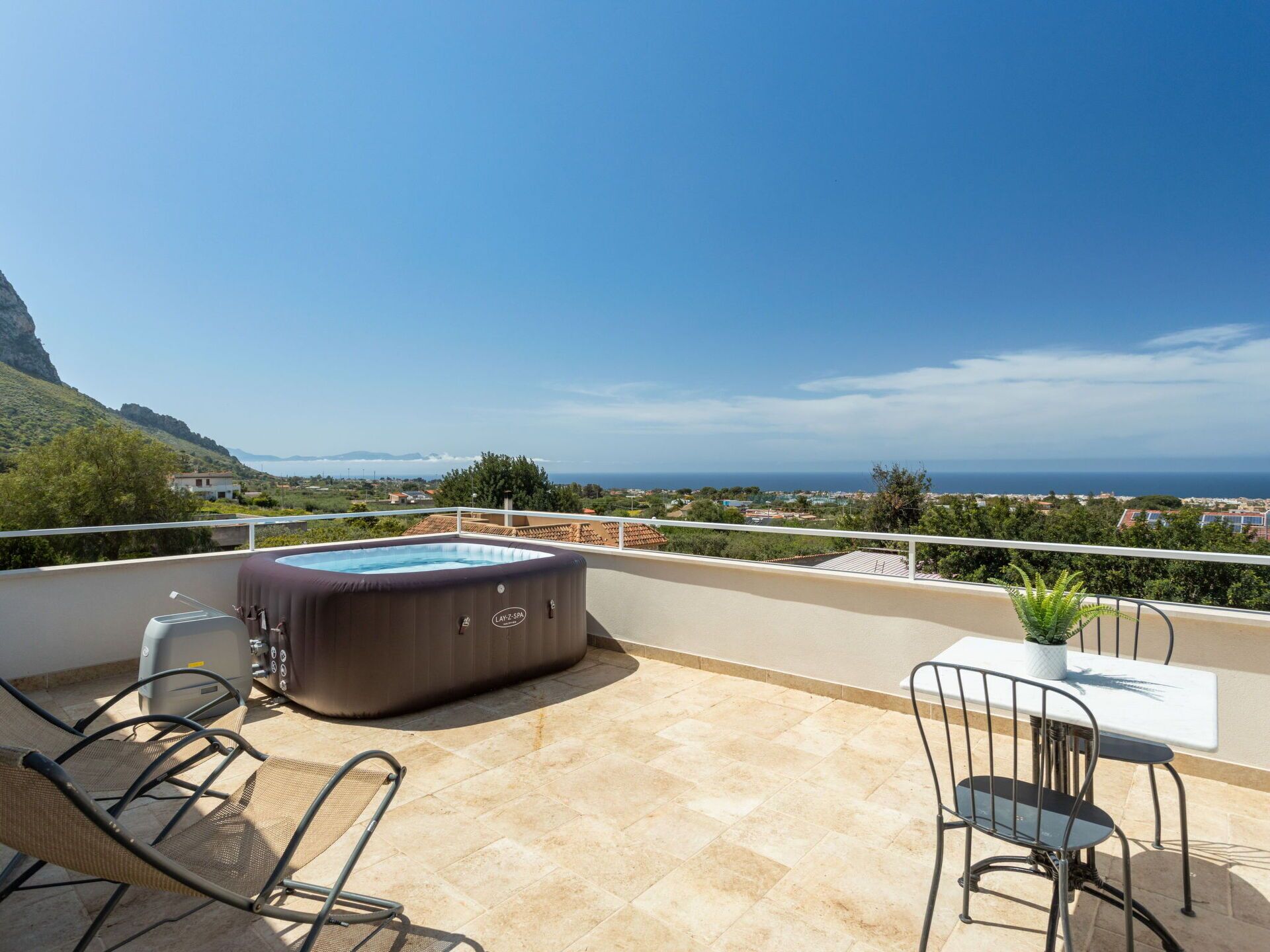 A rooftop terrace with a hot tub, two chairs, a table, and a view of the sea and hills.