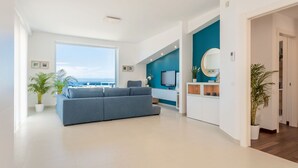 Ein modernes Wohnzimmer mit einem großen grauen Sofa, einem Fernseher und Blick aufs Meer.