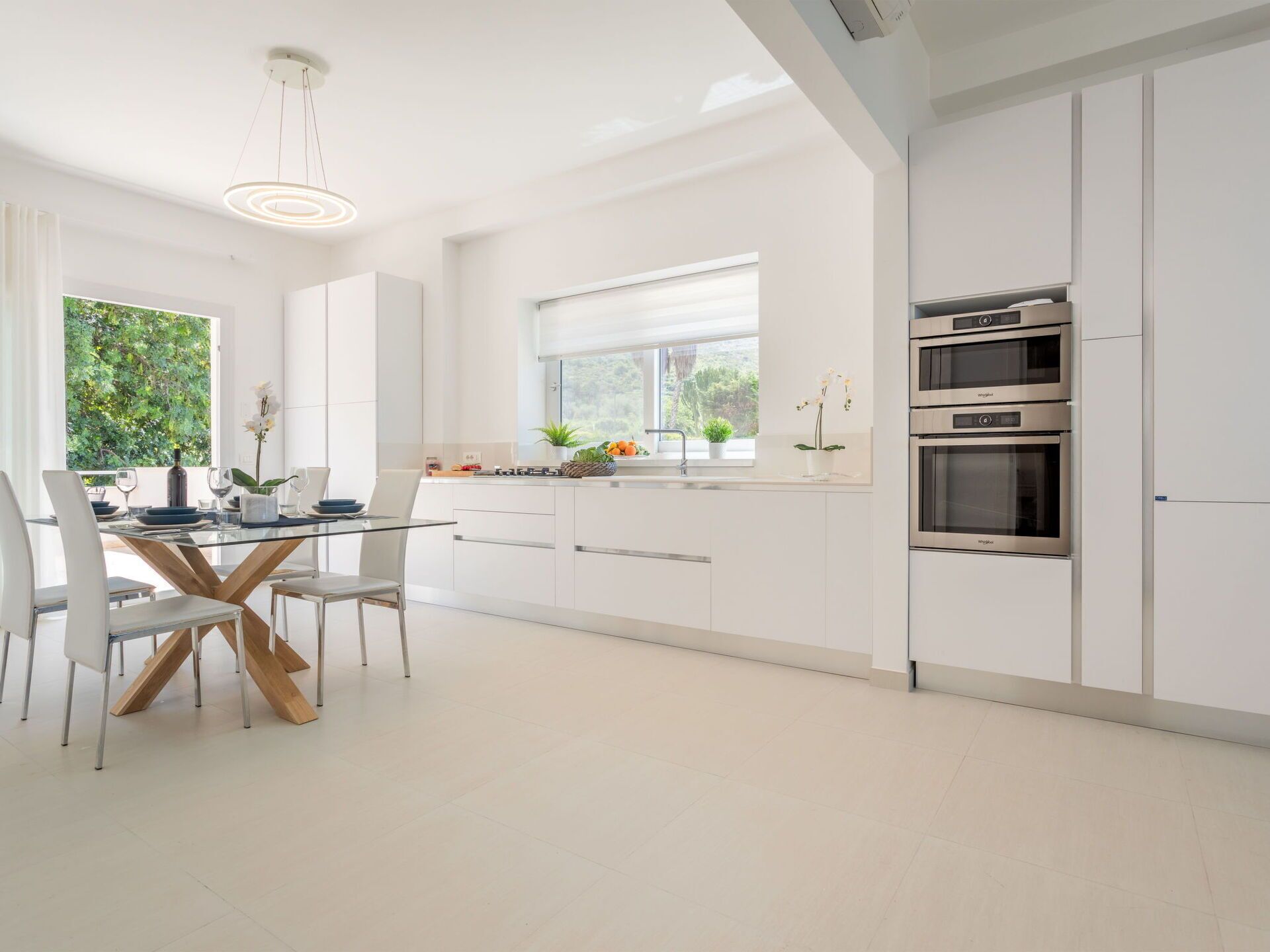 Une cuisine moderne avec des meubles blancs, un coin repas doté d’une table en verre et une vue sur la verdure à l’extérieur.