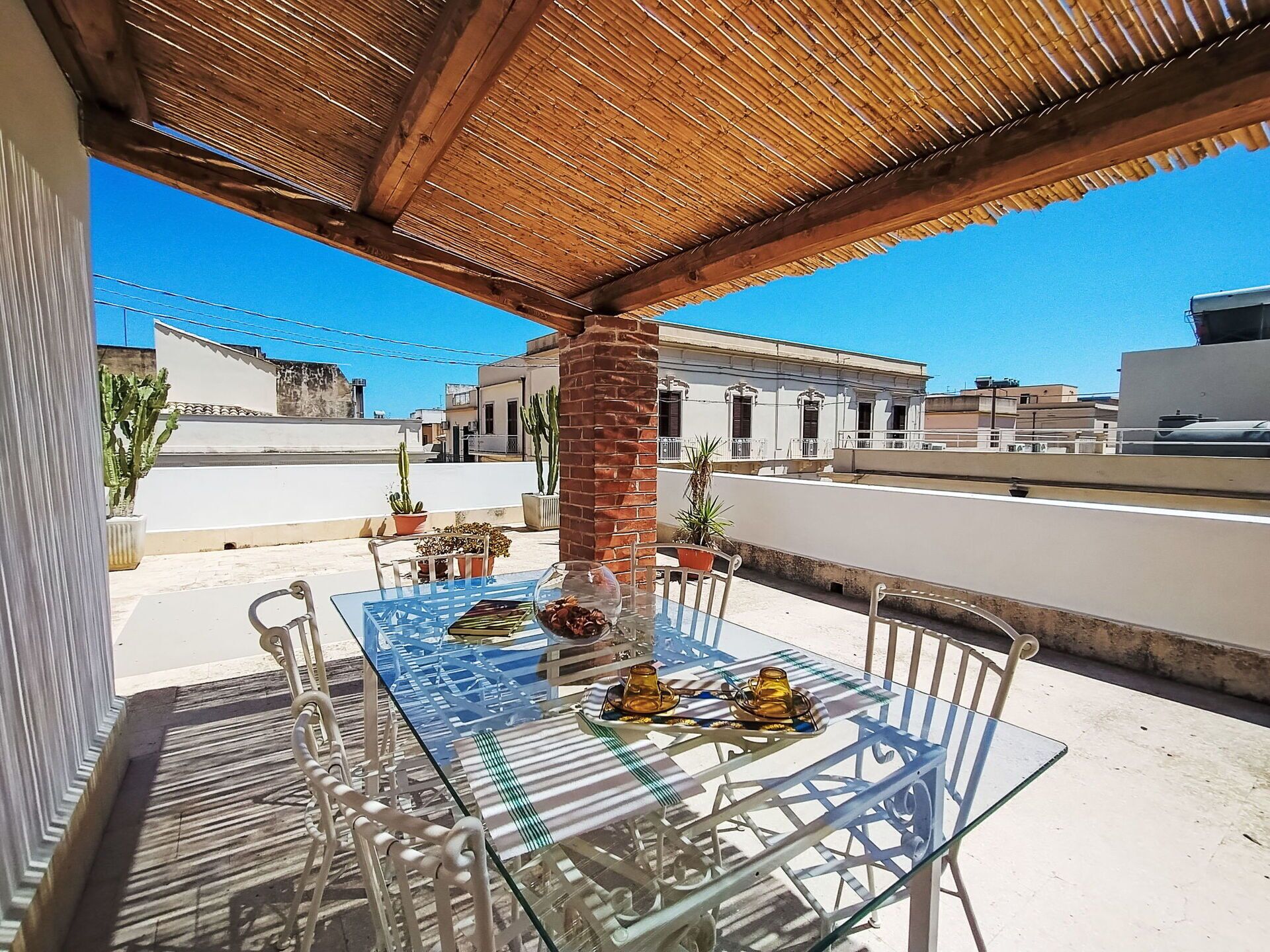 Une table à plateau en verre, des chaises, un pilier en briques et des plantes en pot sur une terrasse sur le toit.