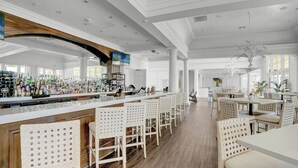 Een ruime bar met een lange toonbank, witte barstoelen en een divers aanbod aan flessen en glazen.