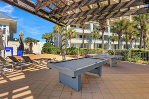 A pool table and lounge chairs in an outdoor area with a view of a multi-story building and palm trees. - The Beach Club 104C by Liquid Life (Gulf Shores)