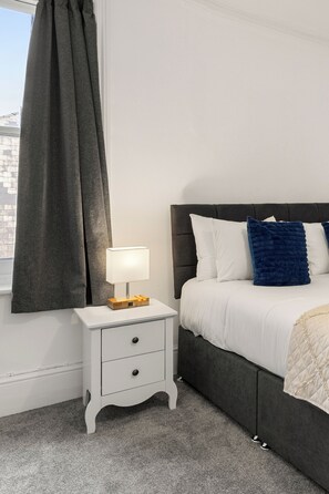 A bedroom with a bed, a nightstand with a lamp, a window with a curtain, and a city view. - Great Titchfield Street 1Bed 1Bath in the heart of London. Newly refurnished (London)