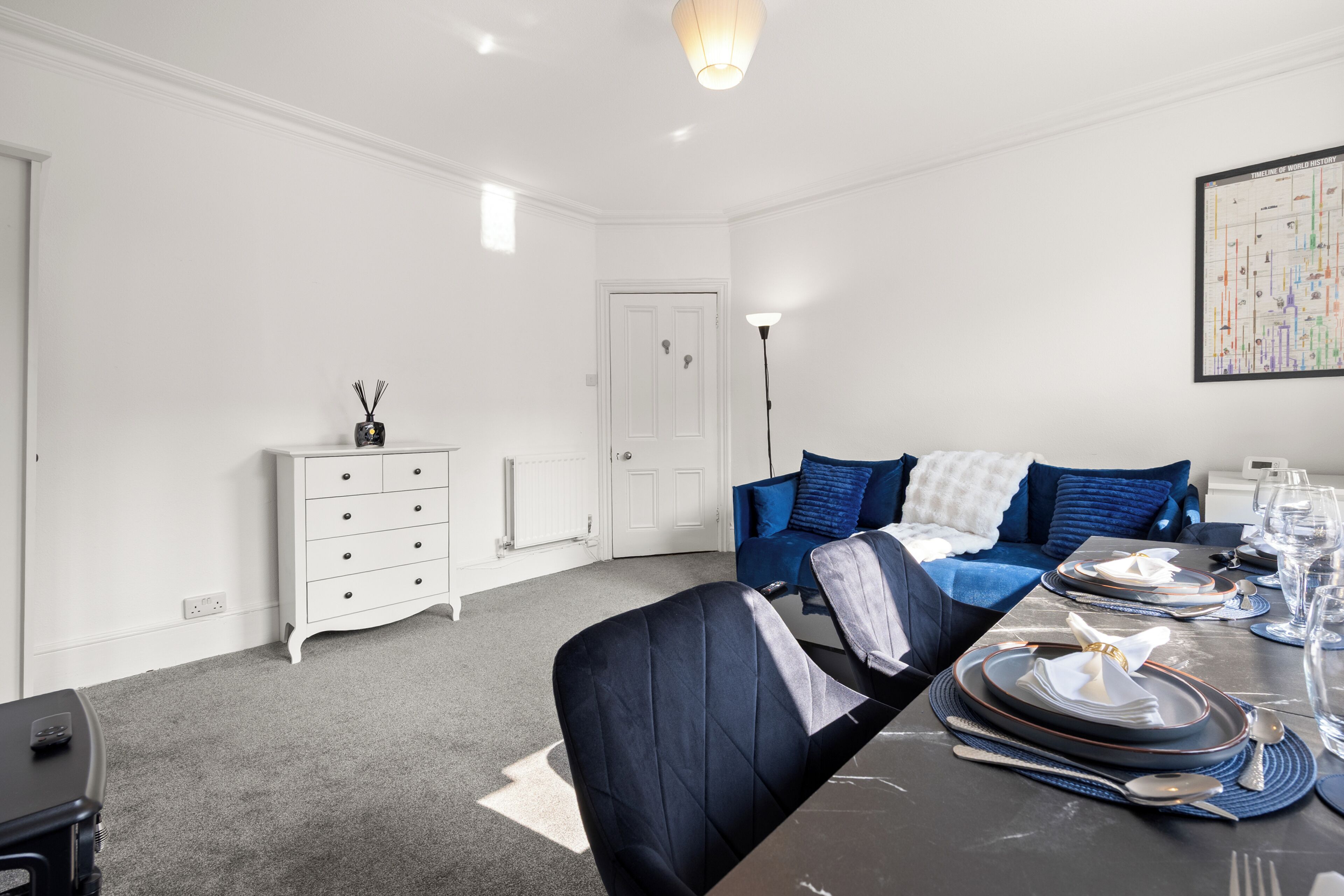 A modern living room with a dining area, white walls, a blue sofa, and a dining table set for a meal.