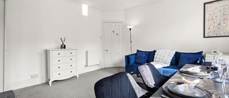 A modern living room with a dining area, white walls, a blue sofa, and a dining table set for a meal.