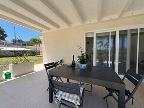 Eine überdachte Terrasse mit Tisch, Stühlen, einem Blumentopf und einer Weinflasche.