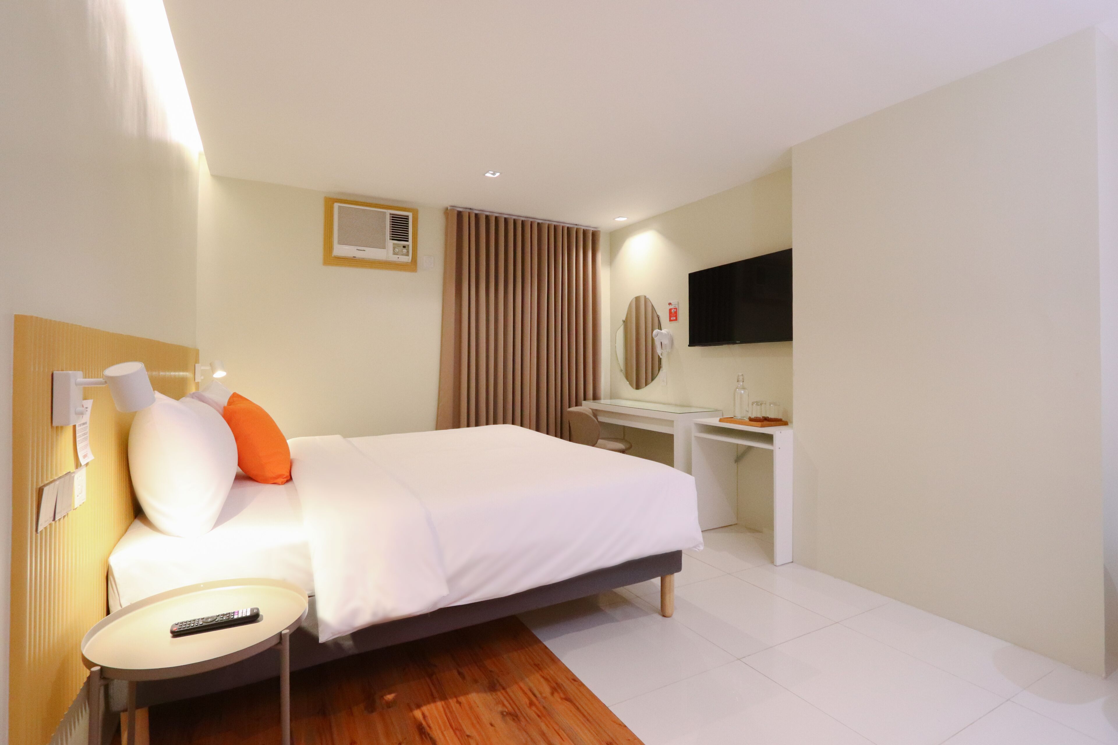 Habitación de hotel con cama, mesita de noche, lámpara, control remoto, escritorio con silla, televisor y espejo.