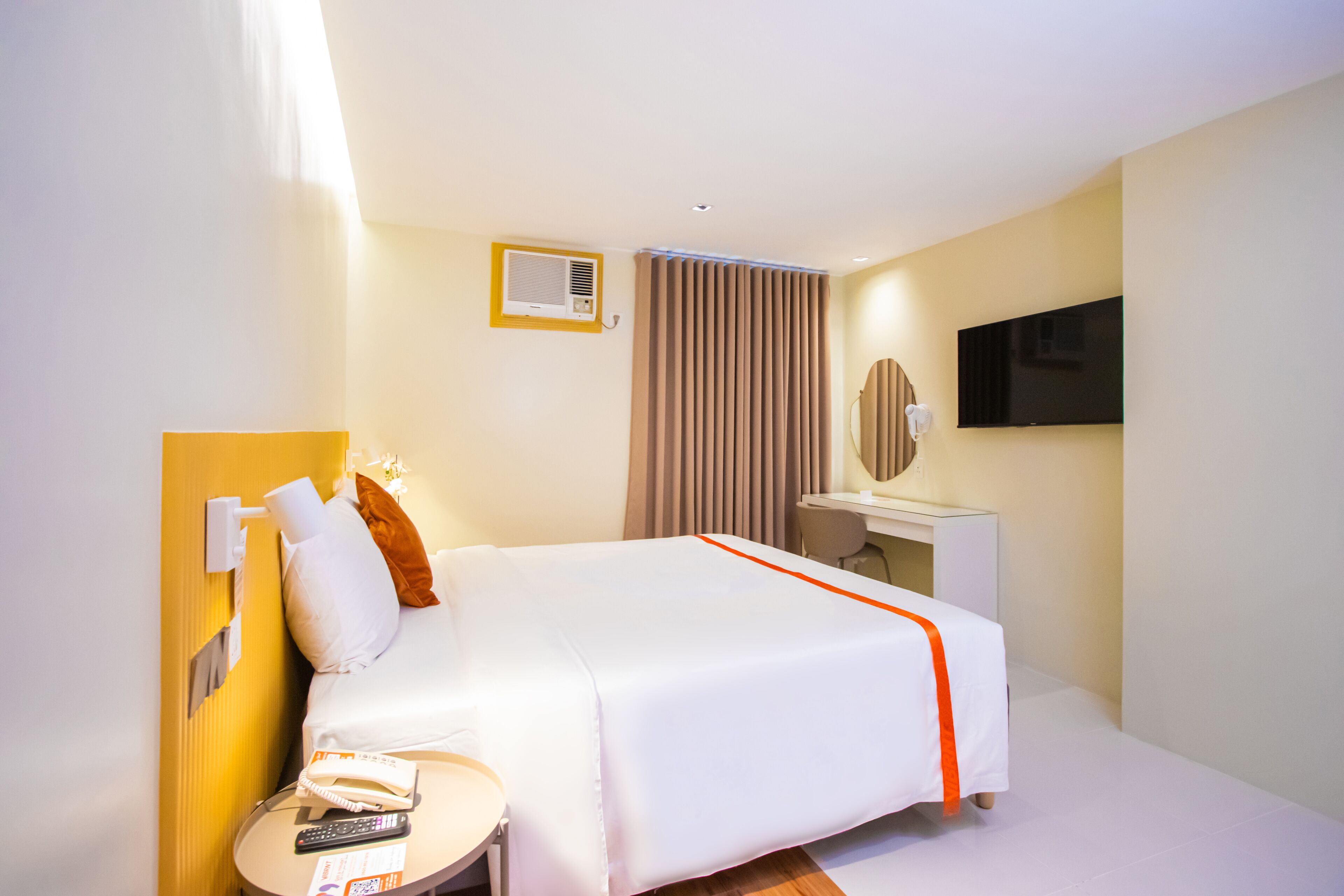 A hotel room with a bed, bedside table, lamp, mirror, TV, and curtains.