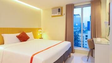 A hotel room with a bed, a desk, a chair, a window with curtains, and an air conditioning unit.