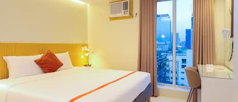 A hotel room with a bed, a desk, a chair, a window with curtains, and an air conditioning unit.