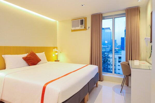 Habitación de hotel con cama, escritorio, silla, ventana con cortinas y aire acondicionado.