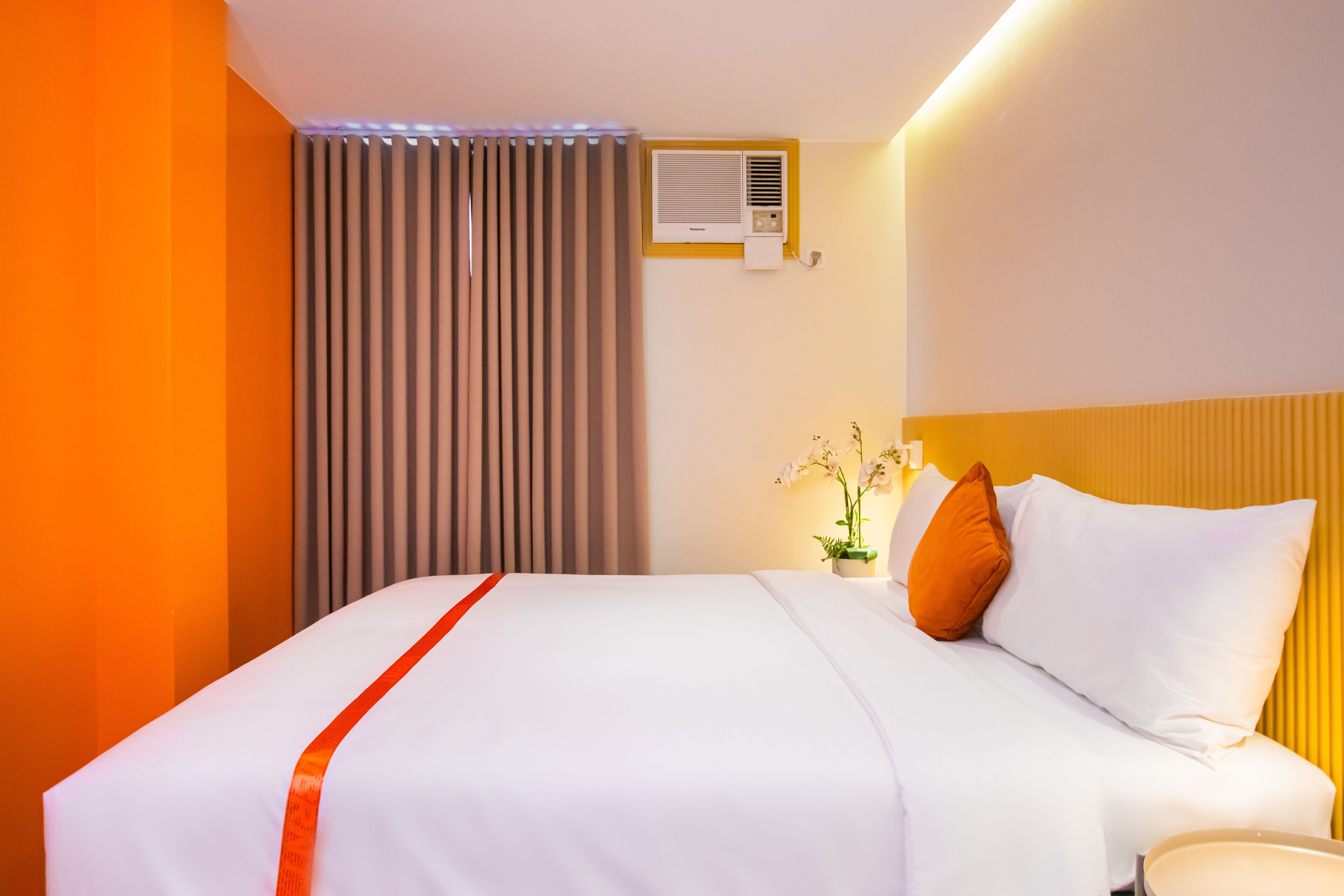 A hotel room with a bed, curtains, an air conditioning unit, a nightstand with a lamp, and a potted plant.