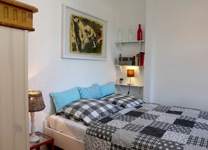 A bedroom with a bed, pillows, a lamp, a wall-mounted shelf with books and bottles, and a framed picture.