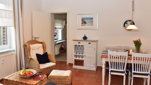 Une pièce chaleureuse avec un fauteuil en osier, une table à manger avec des chaises, un buffet contenant des verres à vin et un tableau accroché au mur.