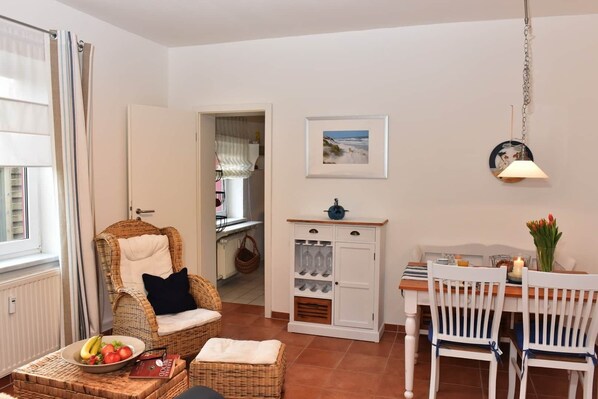 A cozy room with a wicker chair, a dining table with chairs, a cabinet with wine glasses, and a picture on the wall.