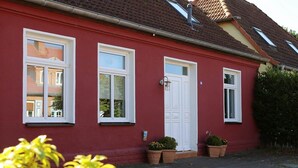 Un bâtiment rouge avec des fenêtres à encadrement blanc, une porte blanche et des plantes en pot à l’entrée.