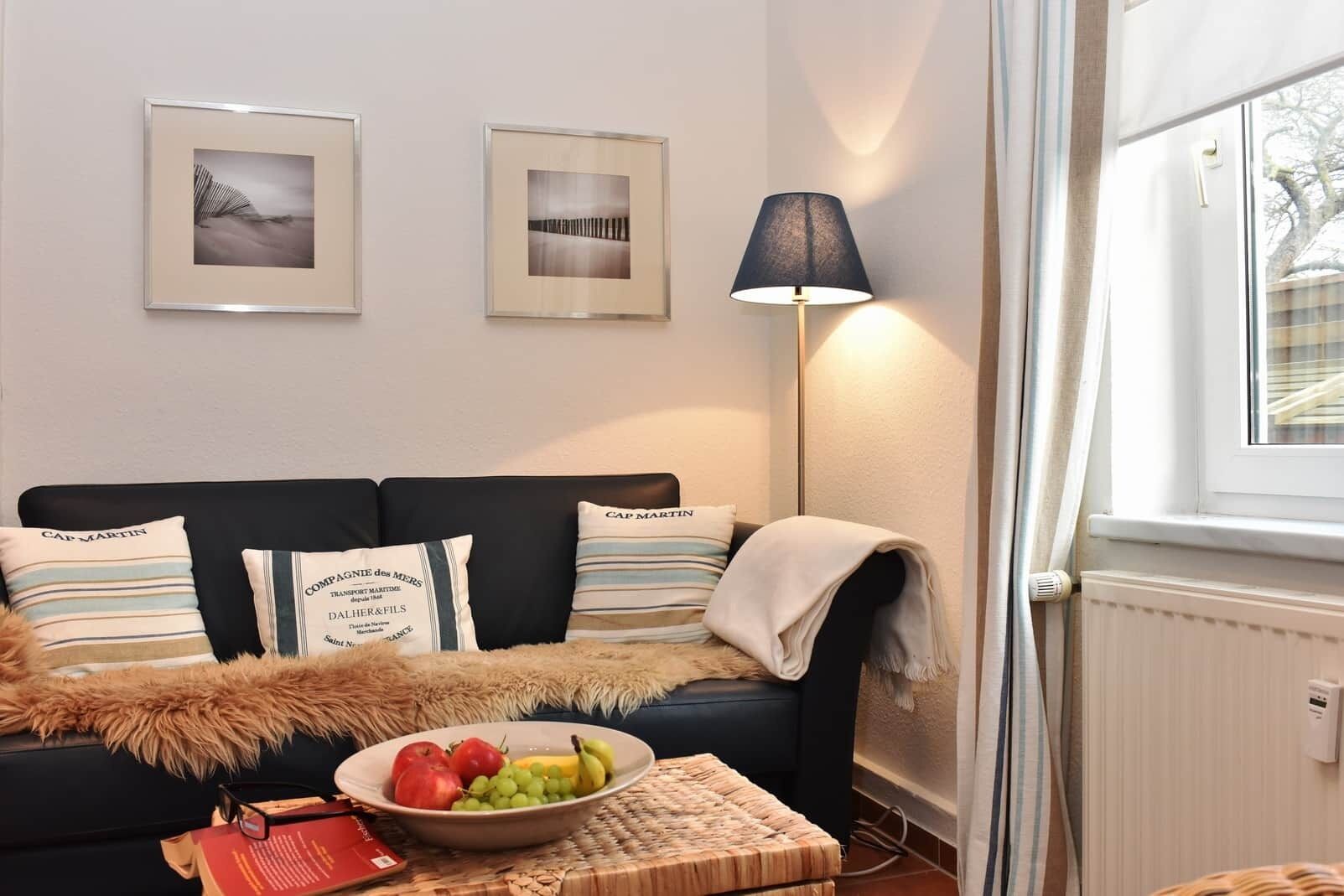 Un salon avec un canapé, deux tableaux encadrés, une lampe, un saladier de fruits et une fenêtre.